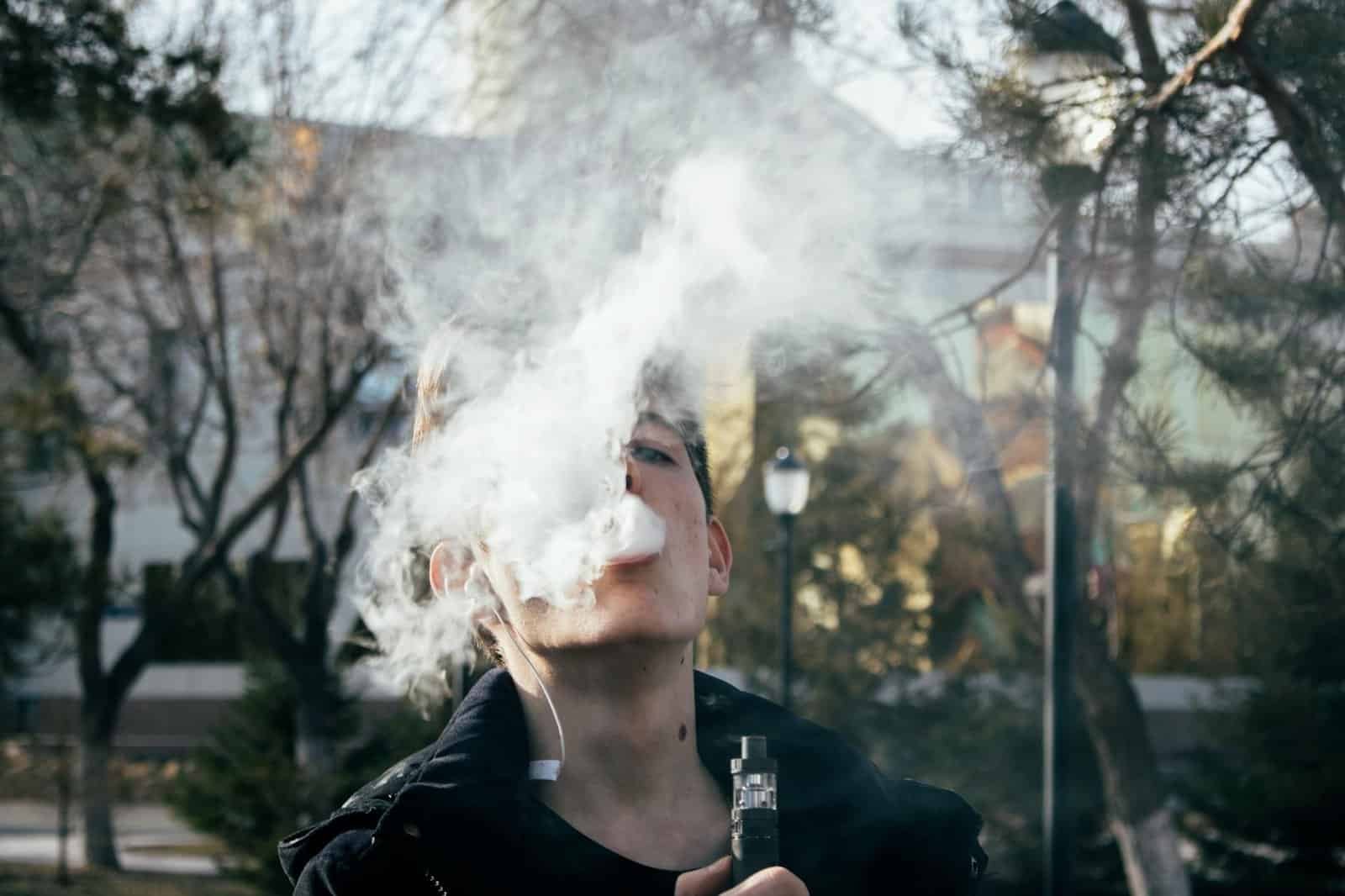 what is vaping in dubai - Portrait of a young man vaping in a park, releasing clouds of vapor on a clear day.
