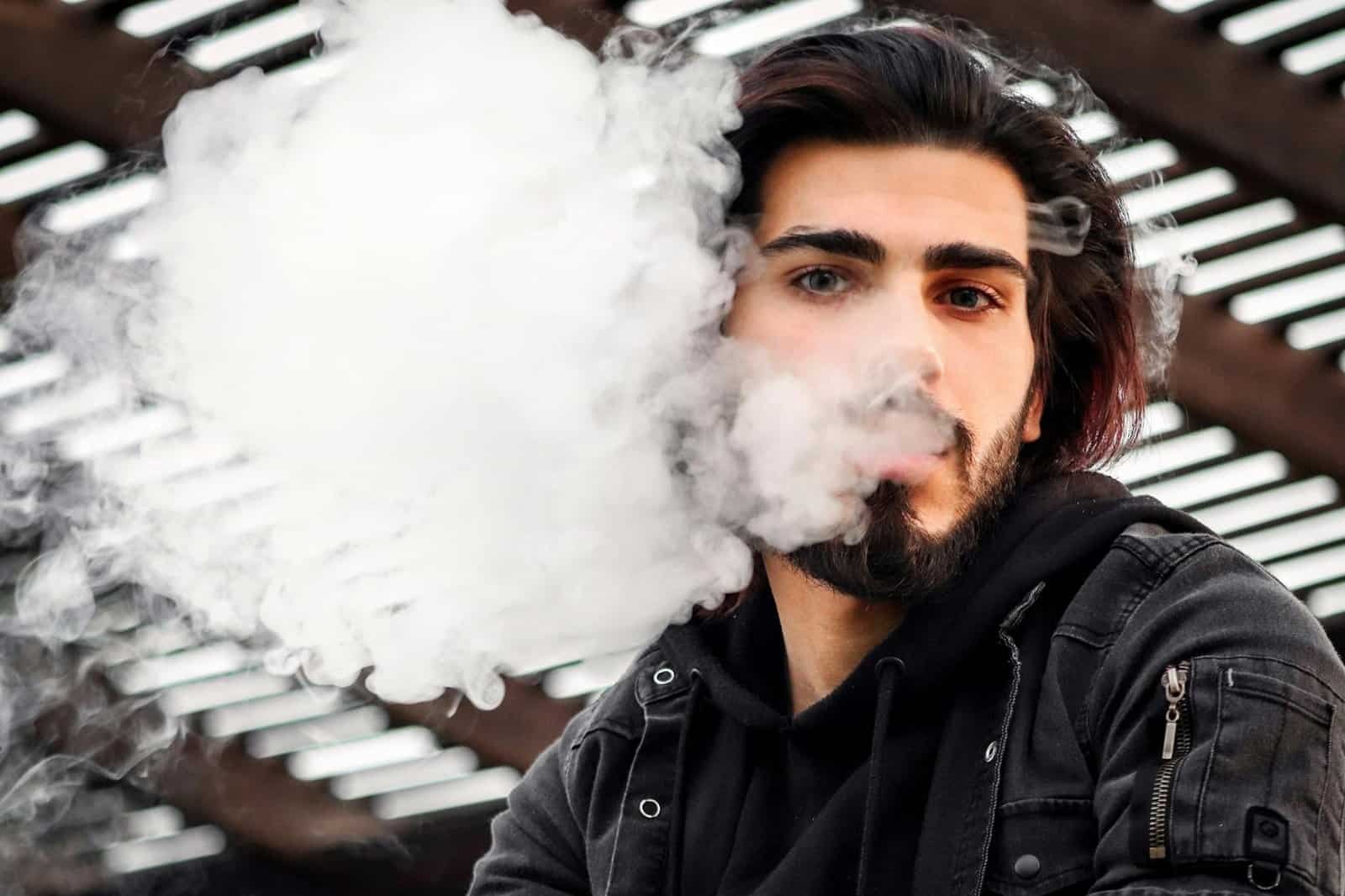 what is vaping in dubai - Man with a beard vaping outdoors, releasing smoke clouds in Dubai.